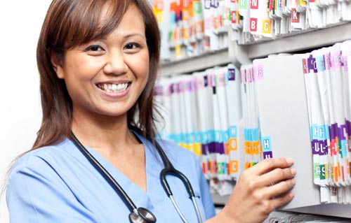 A smiling female nurse in front of a healthcare providers shelf.